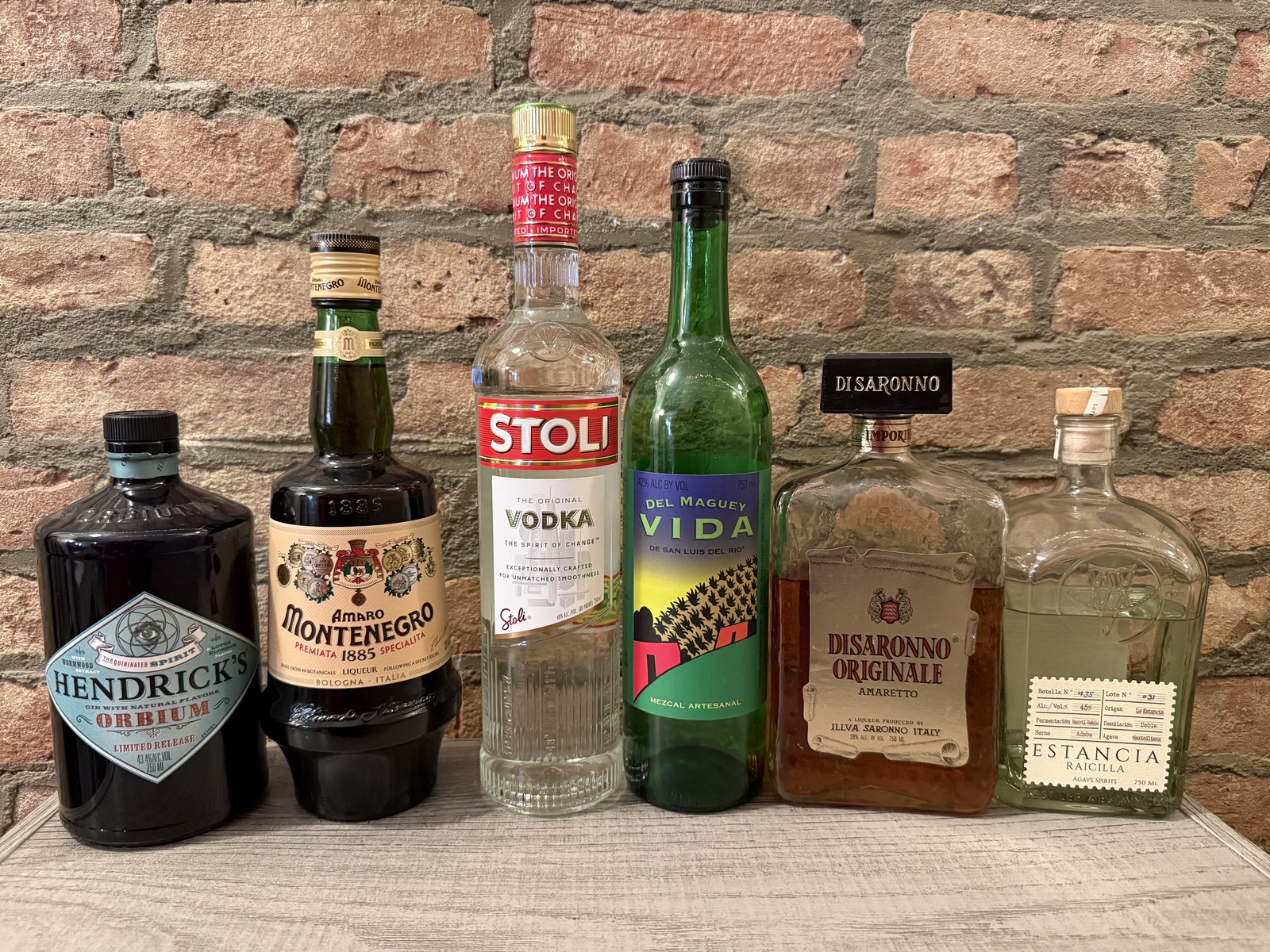 Photograph of six real bottles lined up on a bar: Hendrick's Orbium gin, Amaro Montenegro, Stoli vodka, Del Maguey Vida mezcal, Disaronno amaretto, and Estancia Raicilla