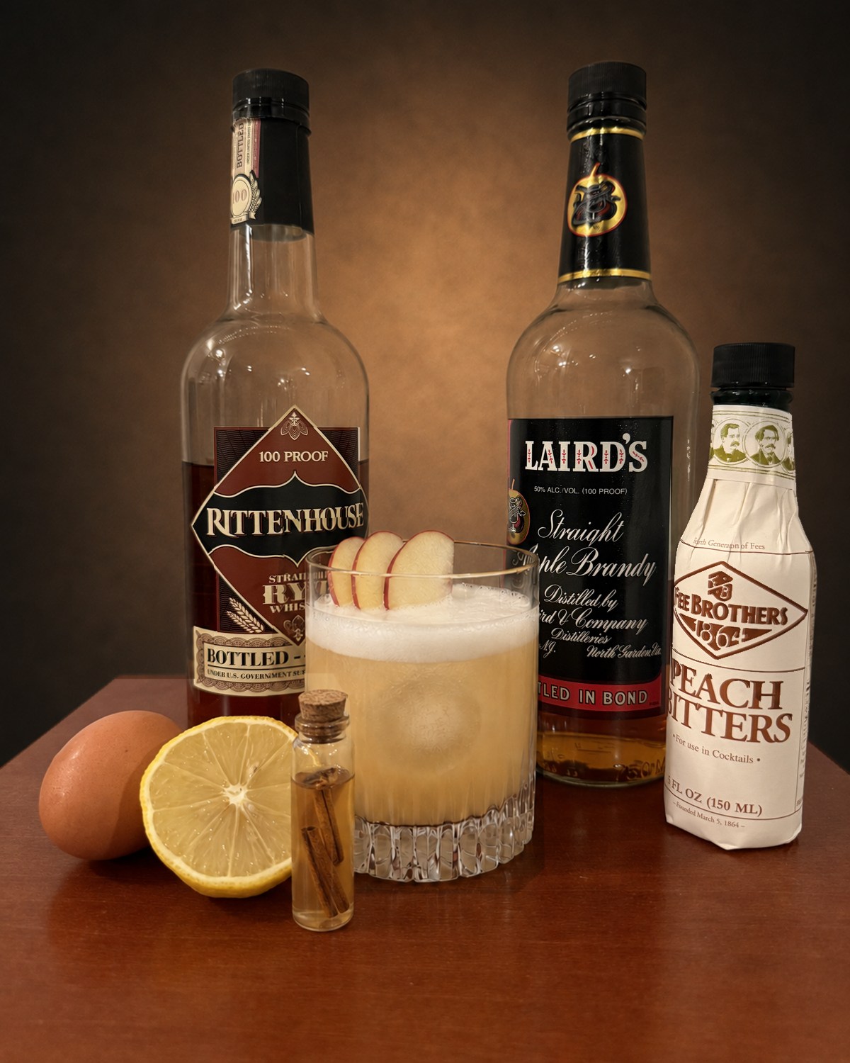 A Big Apple Sour in a rocks glass with a thick egg white foam cap and three fanned apple slices on the rim, flanked by bottles of Rittenhouse Rye Bottled in Bond, Laird's Straight Apple Brandy Bottled in Bond, and Fee Brothers Peach Bitters, with a brown egg, a halved lemon, and a corked vial of cinnamon syrup on the table