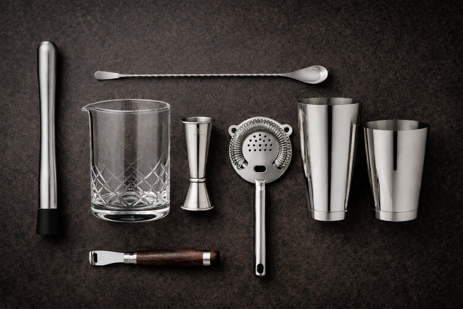 A flat lay of bar tools on a dark surface including mixing glass, bar spoon and jigger