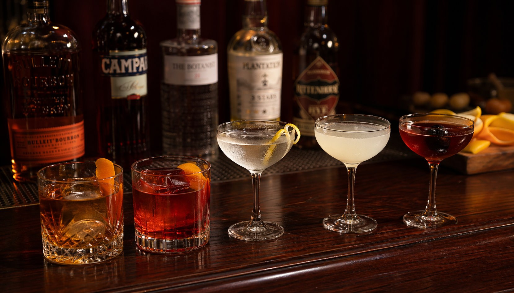 Five classic cocktails arranged in a row on a dark mahogany bar, each in its appropriate glass, with bottles and a citrus peel in the background, warm side lighting