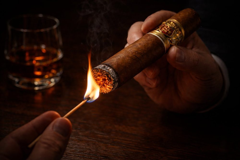 Hands lighting a premium cigar with a match, whiskey glass in background