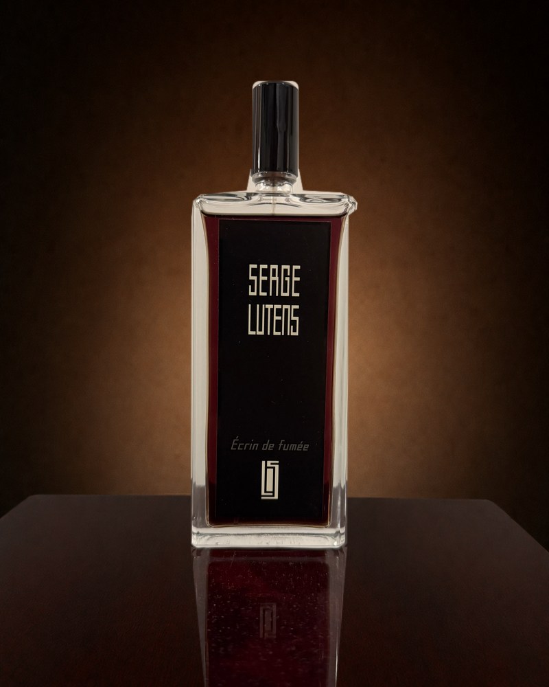 The Serge Lutens Ecrin de Fumee bottle, clear rectangular glass holding deep burgundy liquid, with a black label and a black cap, standing on a dark polished surface against a warm brown background